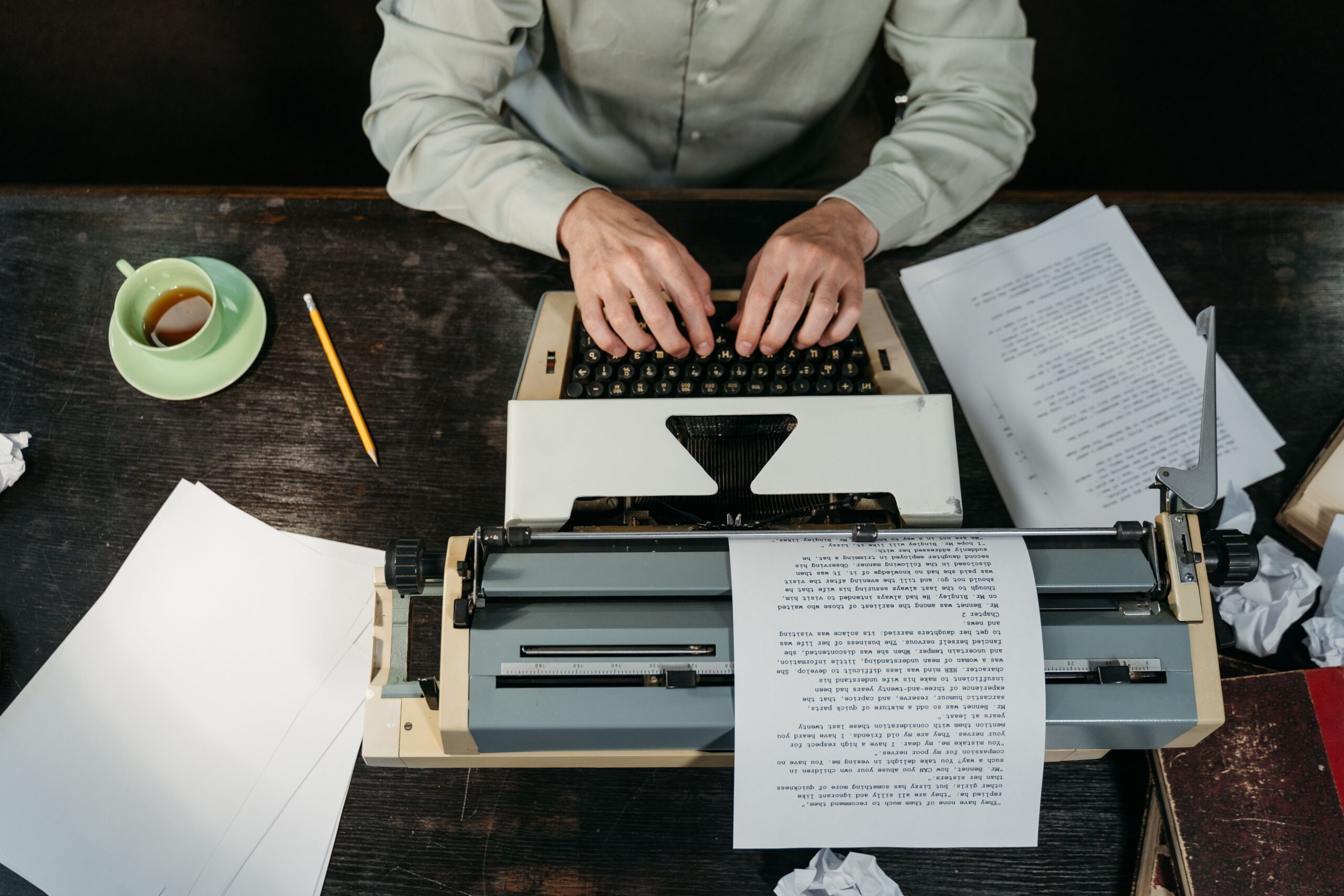 pexels photo 9572418 9572418 Hands typing on a vintage typewriter with papers and a coffee cup in view.